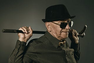 Stylish older man in hat and sunglasses holding cane across shoulders
