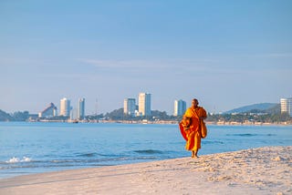 An orange-clad monk walking toward the camera on the beach, with sparse buildings on the horizon under a vast blue sky.