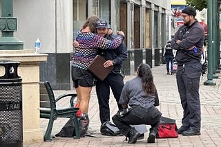 On a city sidewalk in Oakland, CA, a MACRO program responder kneels beside a person on the ground while another responder stands nearby and another person offers a supportive hug.