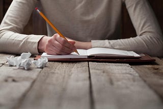 Writing with a pencil in a leather-bound notebook with crumpled drafts on a wooden table.