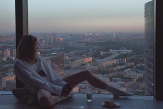 A midlife woman sitting alone beside a large apartment window overlooking a city skyline at dusk, relaxed in a calm modern space, reflecting quietly in peaceful solitude.