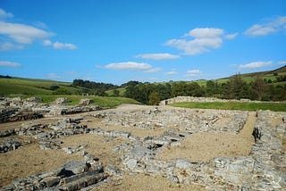 The remnants of rectangular buildings. The remains are low walls, built of greying stone bricks. Light beige gravel paths between the walls are a contrast to the remains. In the background are the hills of the Northumberland Countryside, the sky above is bright, sunny and blue, dotted with white fluffy clouds.