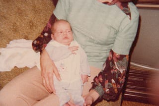 Newborn baby in sleeper with uncomfortable facial expression being held by adoptive mother with 70s style clothing and furninshings