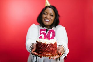 A Black woman with shoulder length straight hair and a party hat wears a white jumper with a pink number 50 on the front. She is smiling and holding a birthday cake, against a red backdrop