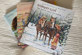 Three Pioneer Girl volumes lay spread on a white blanket. On top is Pioneer Girl: The Path to Fiction, the most recently published volume.