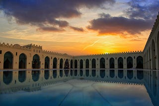 Architecture image, Courtyard bordered by elegant arches, each framed by turquoise drapes. The foreground features a tranquil body of water that perfectly reflects the surrounding structures.