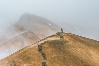 I figure on a hill top. A long path behind them and an even longer path ahead, rising up to bigger hills, shrouded in mist.