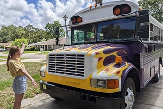 A short 5 window purple bus with flames painted on the front and a redheaded girl arms extended, showing off the bus.
