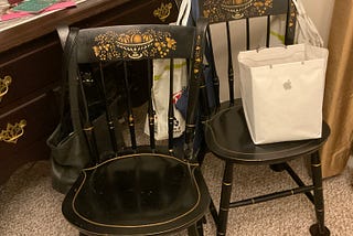 The image shows a room with two black Hitchcock chairs featuring gold stenciling on the backrests. The chairs are positioned in front of a wooden dresser with brass handles, on which there is a tissue box, some papers, and a framed photograph. One chair holds a black leather bag and the other holds a white Apple-branded shopping bag. The decor suggests a personal living space with practical uses for the chairs beyond seating, such as temporary storage or organization.