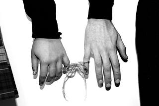 A string tied around fingers with a bow holds two peoples’ hands together.