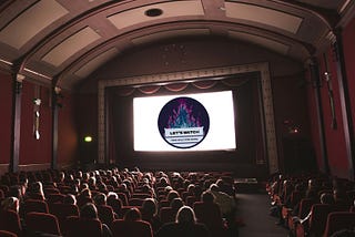 Folks in a movie theater staring at a screen that features the Dumpster Fire logo of While Rome Burns.