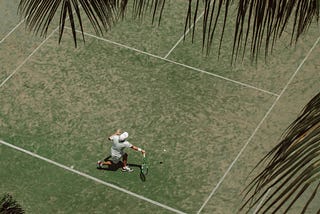 A tennis player, captured from above, serves on a grass court under the shade of palm leaves, evoking a relaxed and tropical atmosphere while highlighting the dynamic movement of the game. Reflecting the perspectie of Tamneek Bhogal on tennis injury prevention and recovery.