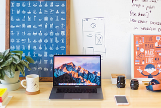 Image of a desk with a laptop, scribbles in a whiteboard and artwork reclined in the wall.