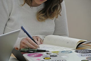 Foto minha com um pullover branco e uma caneta na mão, escrevendo uma lista num caderno aberto em minha frente.