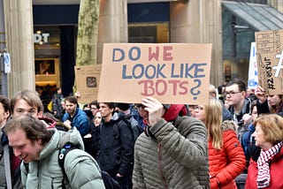 People walking at the demostration, one guy is holding a sign “DO WE LOOK LIKE BOTS?”
