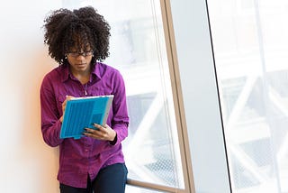 A woman standing indoors with a notebook in hand, jotting down some notes with a focused expression on her face.