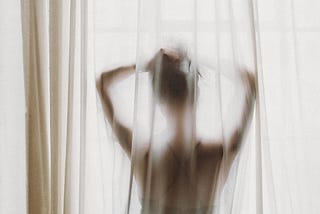 Photo of a woman standing behind a sheer curtain, looking out a window. Her back faces the camera, and the curtain obscures details of her body. Her arms are up, hands holding her hair up.