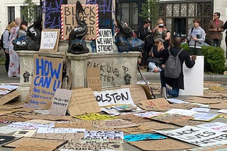 A photograph of the empty pedestal where the statue of Edward Colston once stood, taken on 7 June 2020. The pedestal has graffiti art sprayed on it, and surrounding it are Black Lives Matter and antiracist protest signs. In the background, a small crowd of people are gathered, with a couple of people taking a picture of a Black man beside the empty pedestal with a raised fist and holding a sign in the other hand.