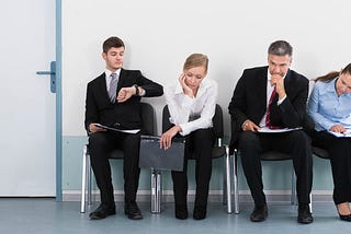 A group of 4 professionally dressed people looking bored sitting outside an office door while waiting to interview.