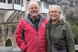 An older couple smiling on a stone bridge in winter in a Balkan town with rocky hills in the background.