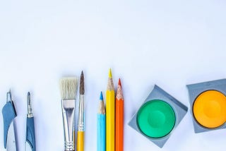 The image shows various art supplies on a white background. Starting on the far left are two metal compasses, continuing are two non-similar size paintbrushes, a set of five colored pencils in bright variegated colors, and a sharpener. On the far right is a circular palette for watercolor paints, and you can see shades of green and orange. Items are diversified across the image with adequate empty white space between them all.