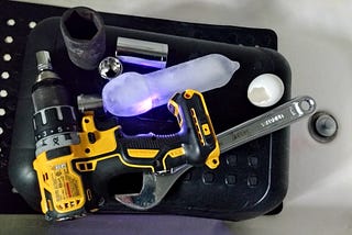 An electric drill with various sockets, an adjustable wrench, an icicle, and an eggshell with a hole in it on top of a black stool in a bath tub. Photo by author.