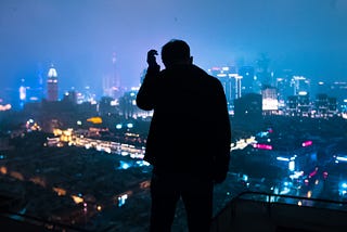 Black silhouette of a man in the center of the image with a city lit up at night behind him.