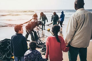 The front part of the image shows a group of people look afar, comprised of an adult and three children. They are holding hands, which suggests that they are a family. At the back, other individuals are seeing picking things from a fishnet at the seashore.