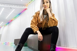 A young woman with long, silver hair sits on a bench looking down at the camera. She’s wearing black jeans and a camel colored jacket with black boots. Strips of neon lights border the room behind her.