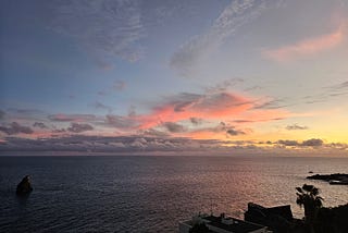 A serene sunset over the ocean with a gradient sky transitioning from soft blue to warm pink hues. Wispy clouds reflect the sun’s colors, and a solitary rock formation rises from the sea in the distance. The coastline to the right is silhouetted with vegetation and buildings against the fading daylight