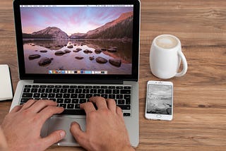 Hands on a laptop keyboard, coffee and phone on desk, lake and mountain image on screen