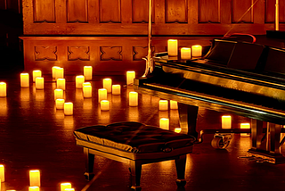 Grand piano and cushioned bench surrounded by glowing candles in a warmly lit, wood-panelled concert hall.