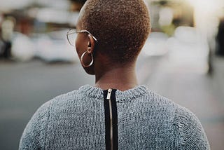 Back view of a black woman looking to the side. She’s wearing a grey sweater with a zipper. One side of her glasses and one of her earrings is showing. Her hair is cut very low and it is brownish.