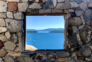 Rustic stone wall with a rectangular hole, looking out onto a vast landscape of blue lake and green woodland in the distance