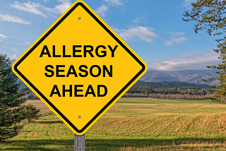 Yellow sign in front of field reading “Allergy Seasons Ahead”