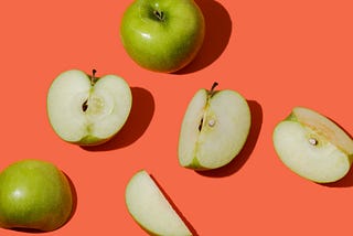 A photograph of several green apples, some cut apart with the cut side facing up, scattered across a bright, almost neon red-pink background.