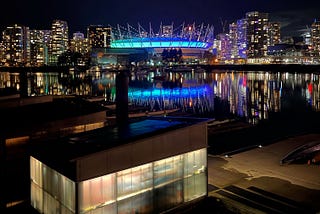 A cityscape at night, showing a lit stadium, hi-rises, and a body of water