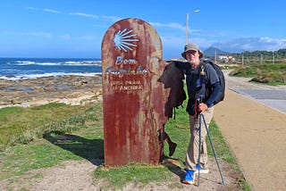 A man with a backpack and hiking poles stands next to a rusted iron sign that says “Bom Caminha. There is a stretch of sand on the right, next to a paved sidewalk, the ocean is on the left side of the photo and a mountain is in the background.