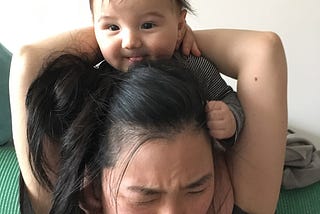 A baby sitting on a mom’s shoulders, gripping her head with a happy expression. The adult appears to be grimacing and struggling.