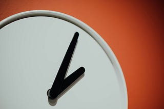 Photo of blank white clock face with black minute and hour hands pointing to roughly 2 p.m. against an orange-red background.