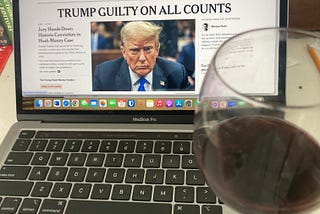 A glass of red wine sits on the edge of a laptop with The New York Times open to a story “TRUMP GUILTY ON ALL COUNTS.” Books, a pencil are also seen on the white desk. (Photo by author.)