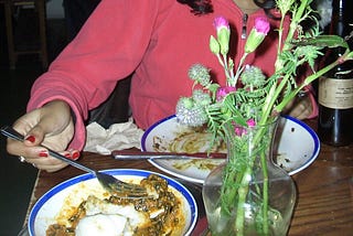 Photo showing a dinner table with two plates of half eaten food. A vase of flowers are on the table and a person opposite in a pink hoodie is reaching for a plate with their fork. The image is cropped so their face is not visible.