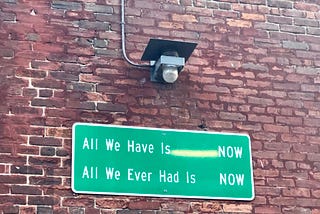 Brick wall with a green sign in the middle that reads, ‘All We Have is NOW. All We Ever Had Is NOW’.