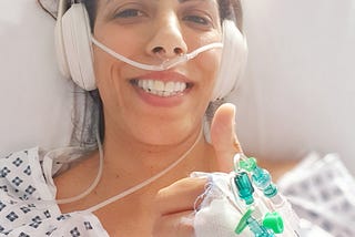 Nav Johal lying in a hospital bed wearing a hospital gown with cannulas in her hand. She is raising a thumbs up and has a big smile on her face. She is also wearing headphones.
