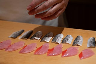 A sushi chef painstakingly prepares small cuts of fish to be used in making the sushi. The delicate slivers of raw fish are carefully laid out on a counter in two rows, only the chef’s hand visible as it hovers above them.