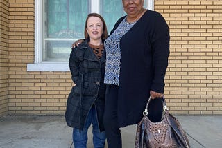 A redhead dwarf woman stands next to an average height African-American woman in front of a yellow brick church