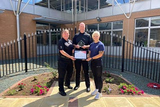 Inspector Harfield (left) standing holding a framed certificate with Ali Booker (right) and Chief Superintendent Spiers (centre) at MDP Headquarters, there are railings and a building in the background