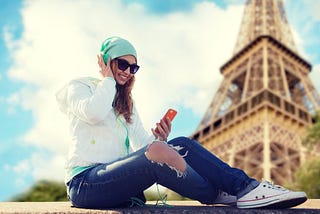 A photo of a woman listening to a podcast on her phone with the Eiffel tower (Paris) in the background.