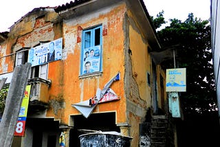 Photo of the house’s facade with hanging election banners. Walls unplastered in several areas.