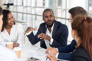 Millennial employees gathered in boardroom for training, black man speaking to Black and white co-workers and corporate team.